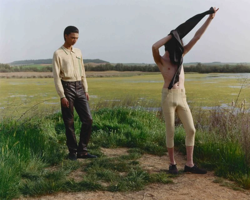 Two men in outdoor setting. One man wearing dark pants, the other shirtless holding a jacket. Marshland and hills in the background.