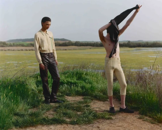 Two men in outdoor setting. One man wearing dark pants, the other shirtless holding a jacket. Marshland and hills in the background.