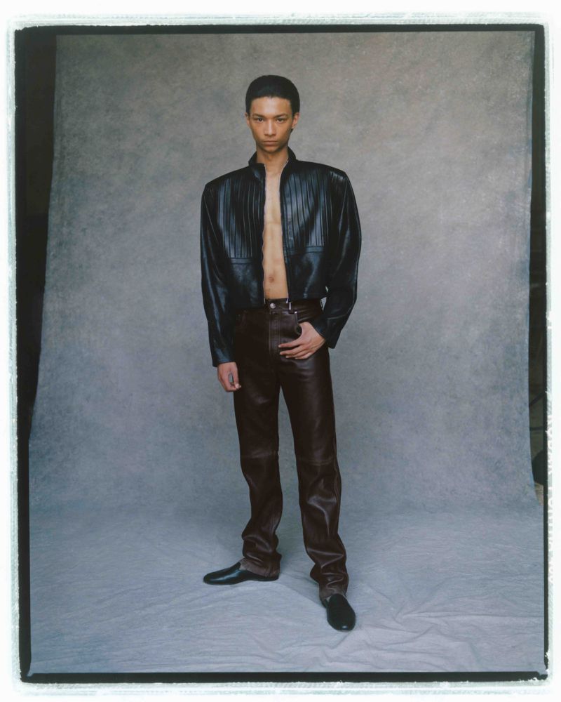 Fashion shot of a young adult male in a black leather jacket and brown leather pants, posing in front of a grey backdrop in a studio.
