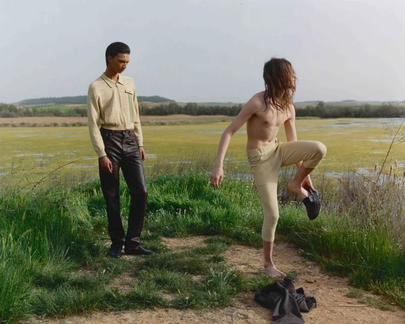 Two people in a grassy field with a body of water in the background. One man is putting on his shoe while the other watches.