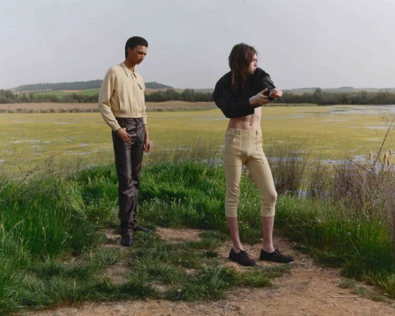 Two people stand in a grassy field near a body of water. One person wears a beige jacket and dark pants, the other adjusts a black jacket.