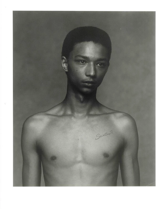 Monochrome studio portrait of a shirtless young Black man with a chest tattoo against a soft textured background.