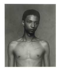 Monochrome studio portrait of a shirtless young Black man with a chest tattoo against a soft textured background.