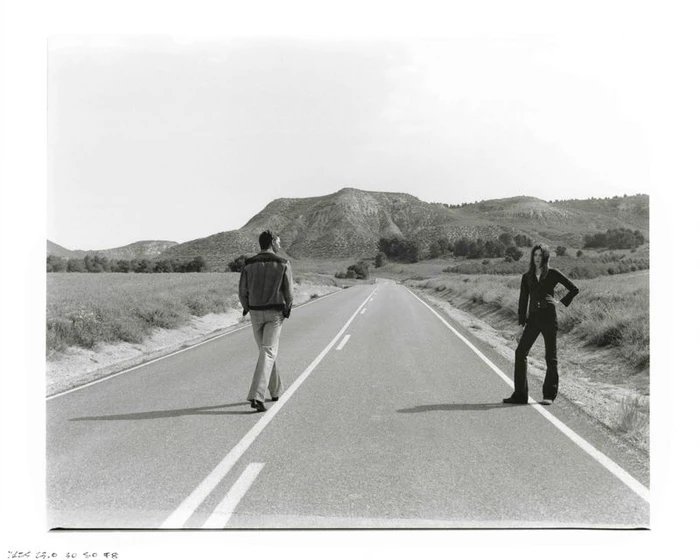 A man walks away along a white road lane line while a woman stands on the road shoulder against a distant mountain backdrop.