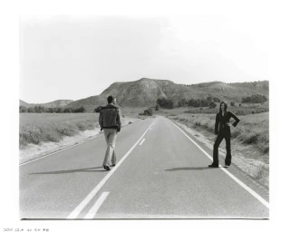 A man walks away along a white road lane line while a woman stands on the road shoulder against a distant mountain backdrop.