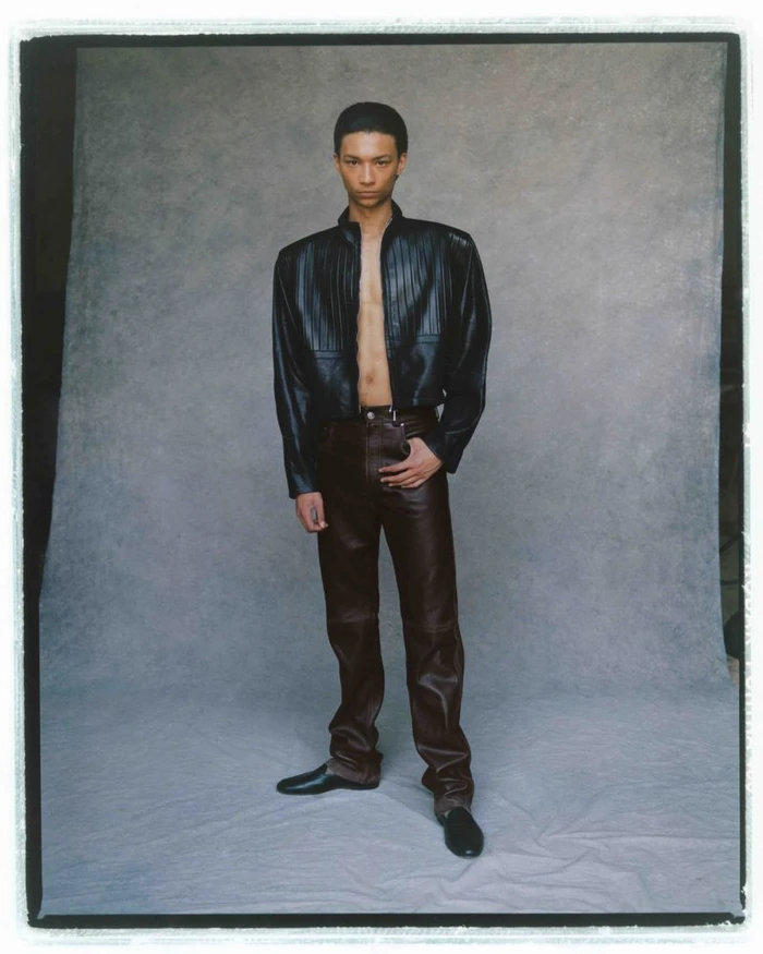 Model posing in a cropped black leather jacket and dark brown leather trousers against a textured grey studio backdrop.