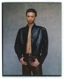 A male model wearing an open black pleated leather jacket and brown leather pants against a textured grey backdrop.