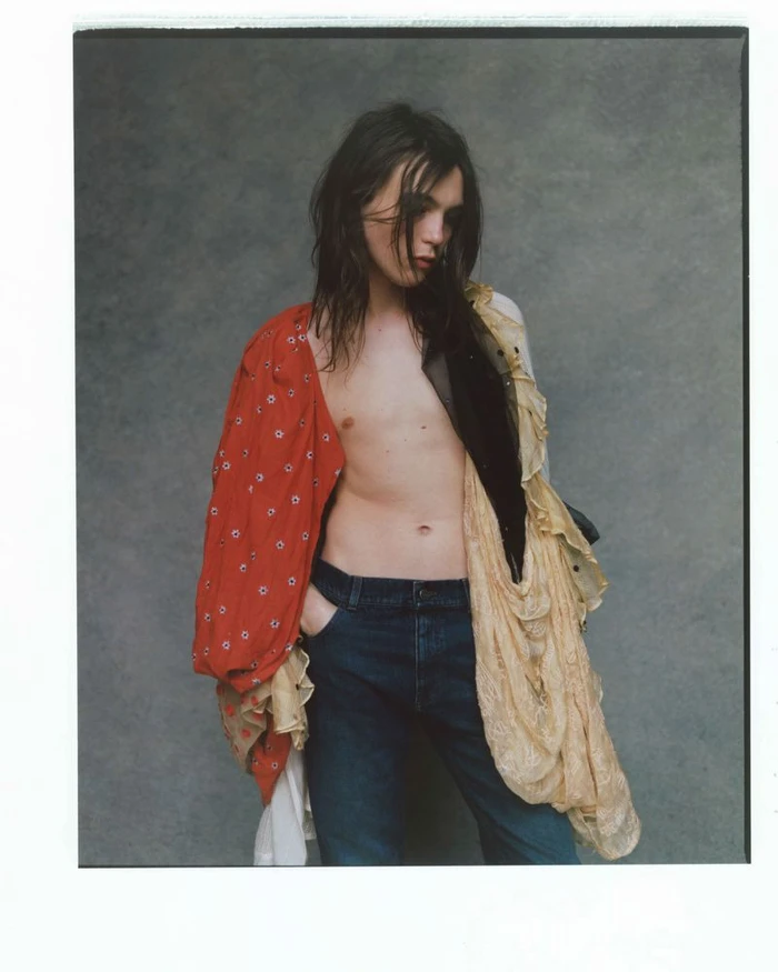Portrait of a young male model with long dark hair wearing unbuttoned layered garments and dark jeans against a textured grey studio wall.