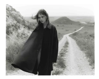 Model wearing an oversized black flared coat standing beside a rural dirt road in a black and white landscape.