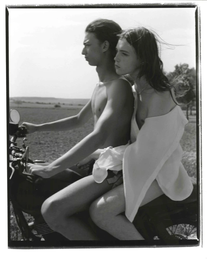 Black and white photo of two models riding a motorcycle through an open rural field.