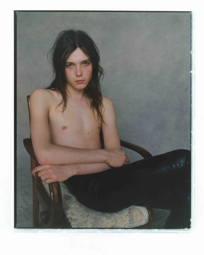 Shirtless young man with long dark hair seated in a vintage armchair wearing black leather pants against a textured grey backdrop.
