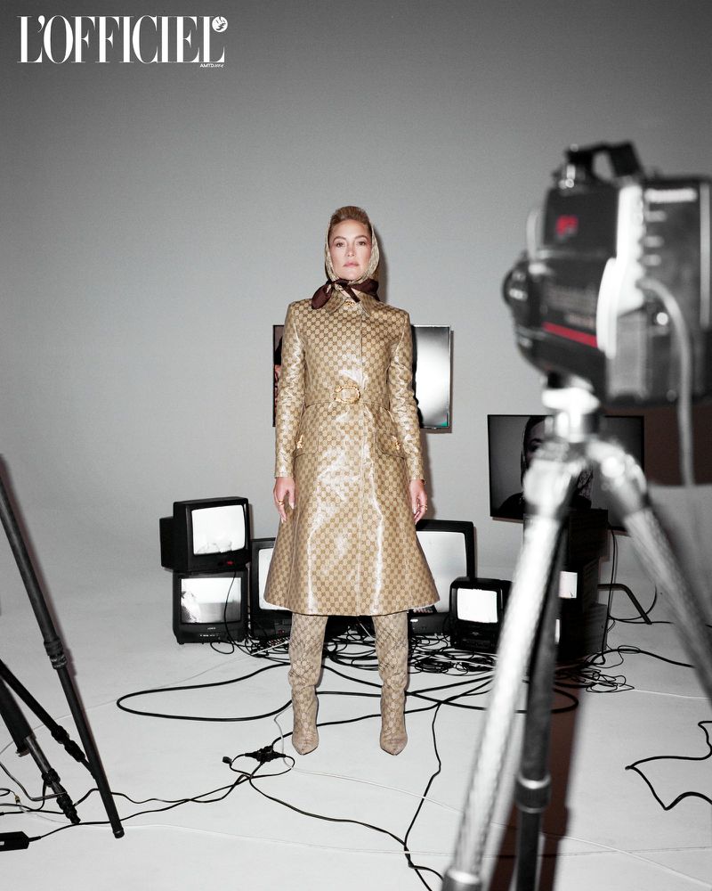 A blonde woman stands in a tan coat and boots against a grey backdrop with vintage TVs and camera equipment in a fashion editorial shot.