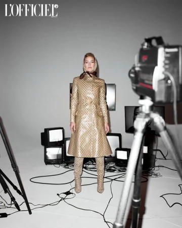 A blonde woman stands in a tan coat and boots against a grey backdrop with vintage TVs and camera equipment in a fashion editorial shot.
