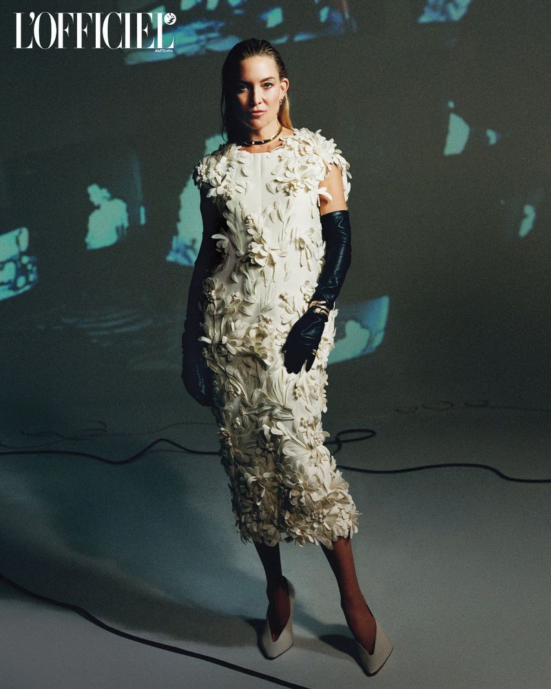 A woman in a white dress stands in front of a projected background in a fashion pose, wearing long black gloves and matching high heels.