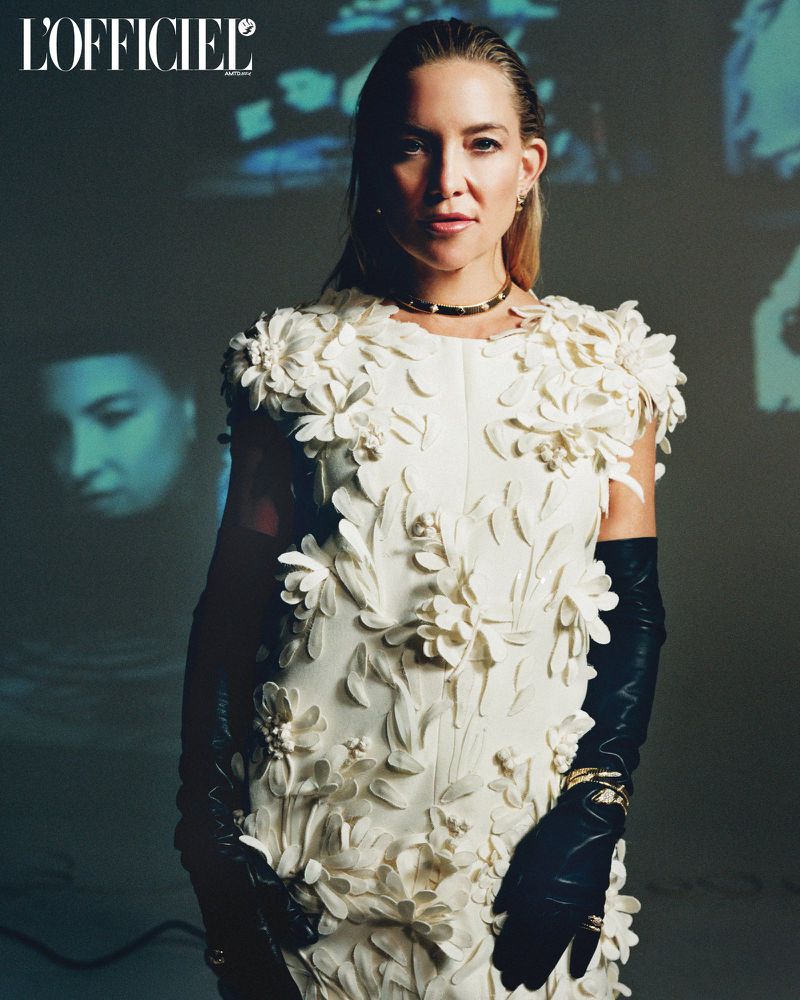 Portrait of Kate Hudson wearing a white floral dress and black leather gloves, posing against a muted background for L'Officiel magazine.