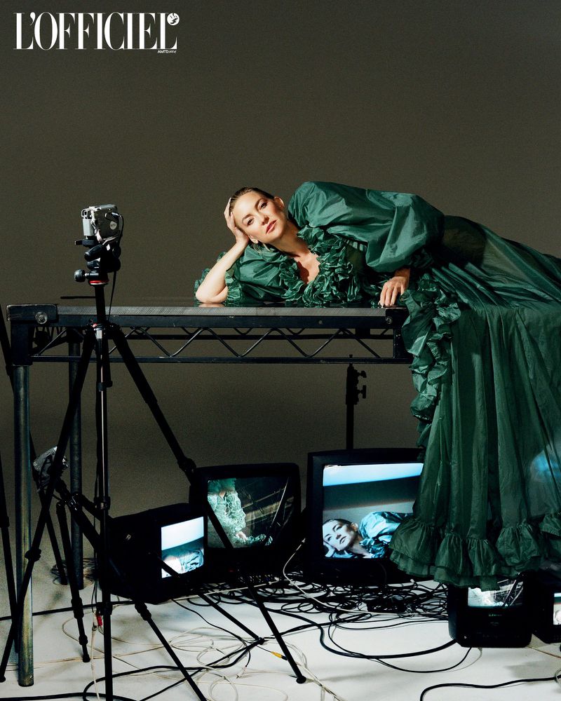 Adult female fashion model in green gown, reclining on table near camera and monitors in photo studio. Editorial fashion photography.