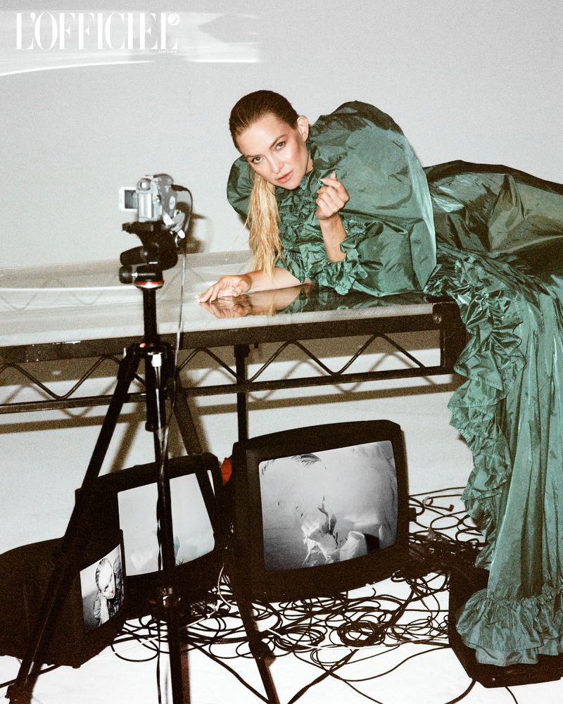 Model in green dress poses on reflective surface with vintage video equipment in a fashion editorial shot for L'Officiel magazine.