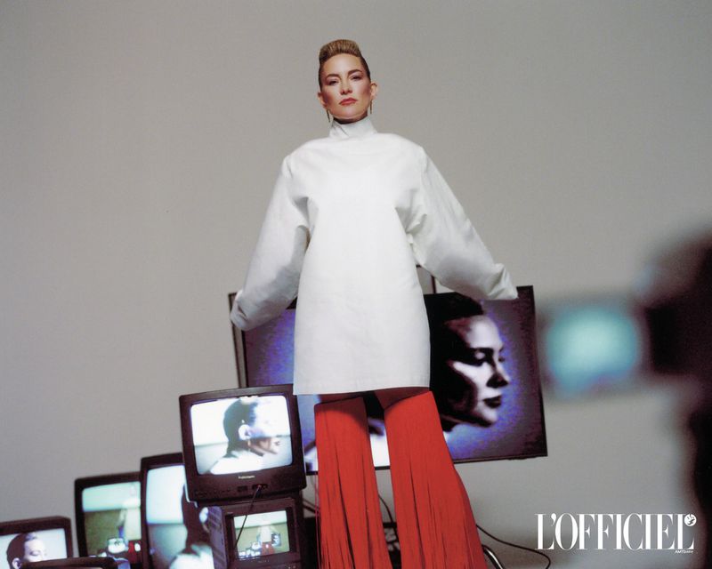 Fashion shot of a woman in a white dress and red pants standing in front of stacked vintage televisions in a brightly lit photo studio.
