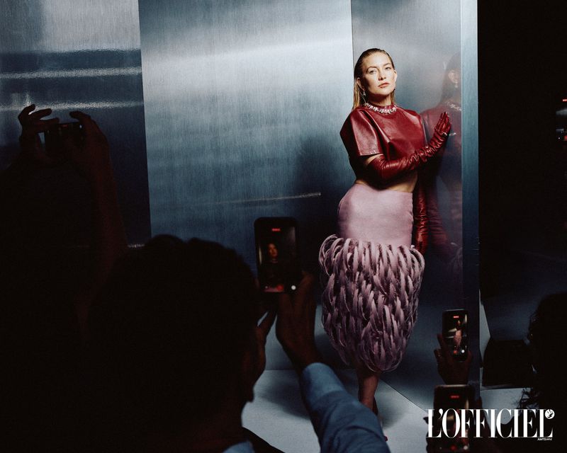 Kate Hudson poses in a red leather top, long red gloves, and a pink feather skirt while photographers take photos with their phones.