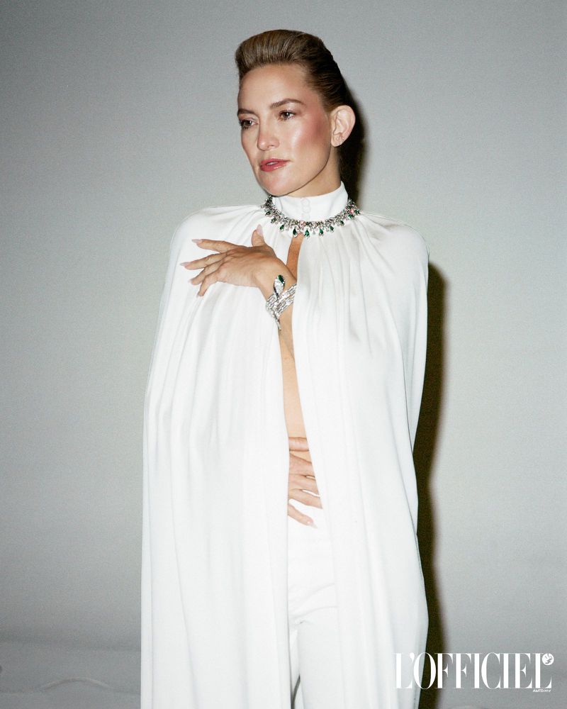 Elegant woman in a white cape and jewelry, posing in front of a light grey background. The image is for L'Officiel magazine.