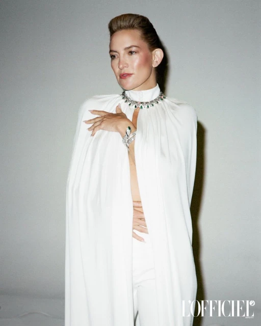 Elegant woman in a white cape and jewelry, posing in front of a light grey background. The image is for L'Officiel magazine.