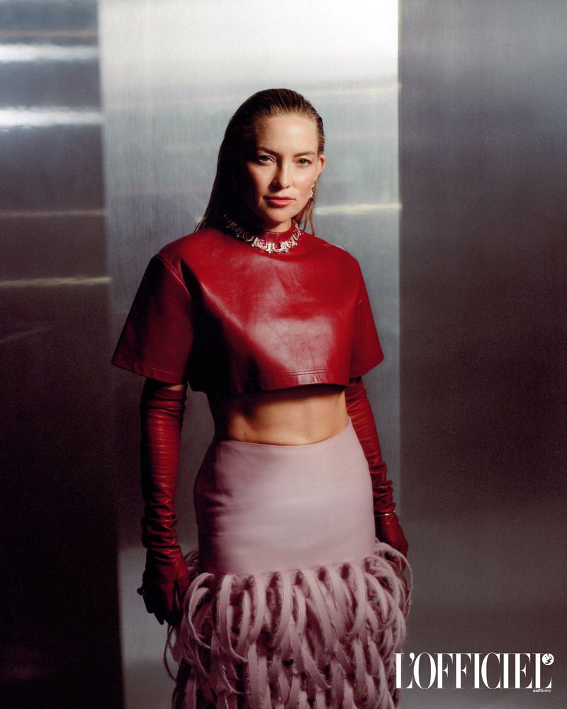 Kate Hudson poses in a red crop top and long gloves paired with a pink feathered skirt against a metallic background in a medium shot.