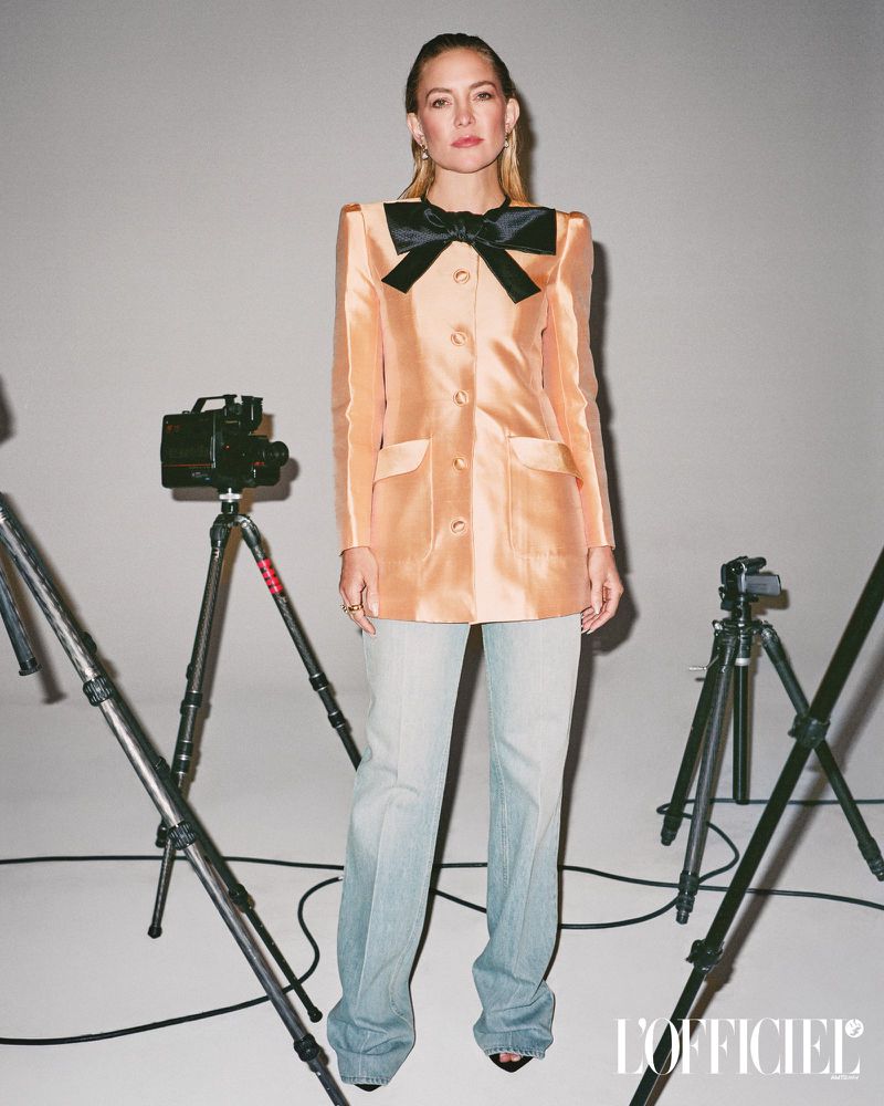 Kate Hudson poses in a peach blazer with a large black bow, light blue jeans, and heels in front of vintage cameras on tripods.
