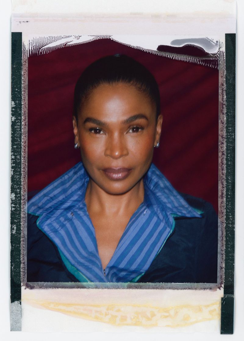 Portrait of a woman with short hair wearing a blue striped collared shirt and dark jacket, photographed in front of a red background.