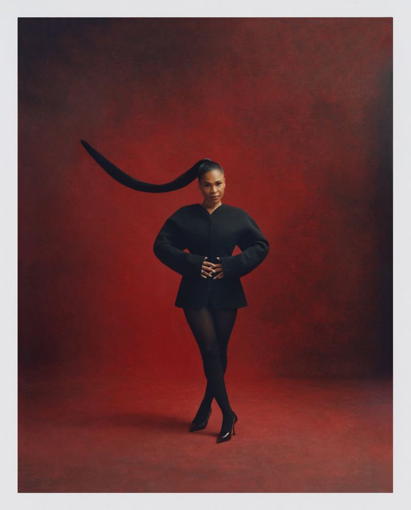 Nia Long wearing a black structured jacket and tights against a deep red background with her long hair in a curved high ponytail
