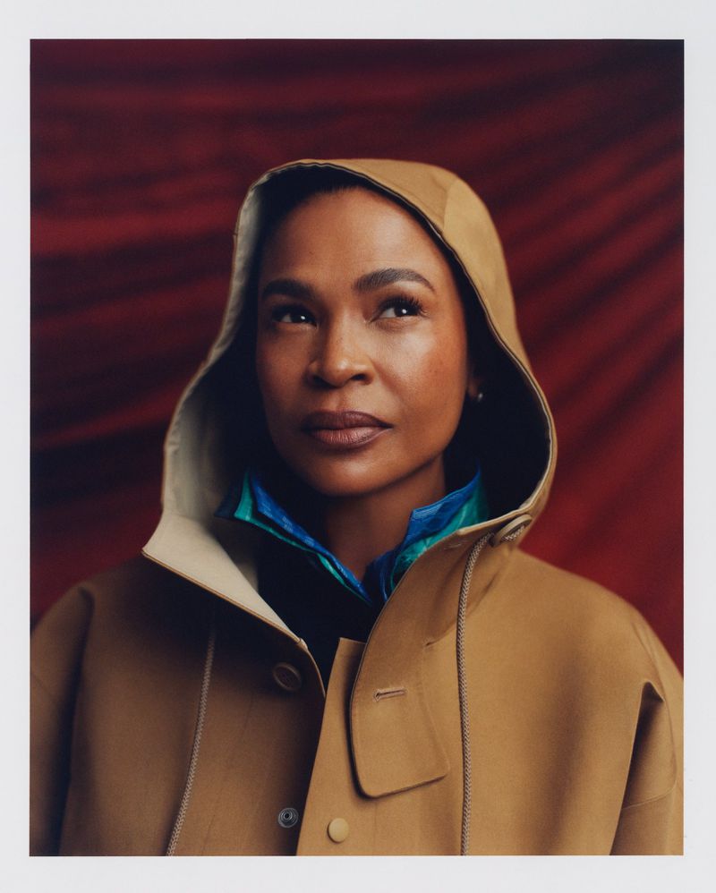 Portrait of an African American woman wearing a tan hooded jacket against a textured red backdrop, looking slightly upwards.