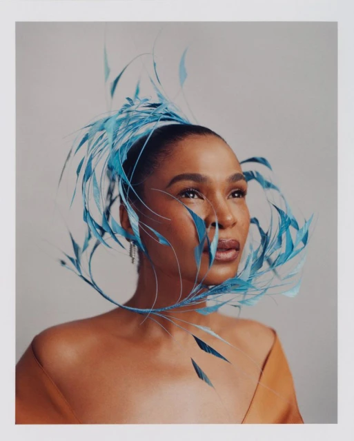 Elegant portrait of a Black woman wearing a blue feathered headpiece, adorned with a strapless dress, gazing upward in a studio setting.