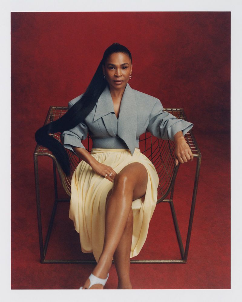 A woman with long, dark hair poses in a wire chair against a red background, wearing a gray jacket and a yellow skirt.