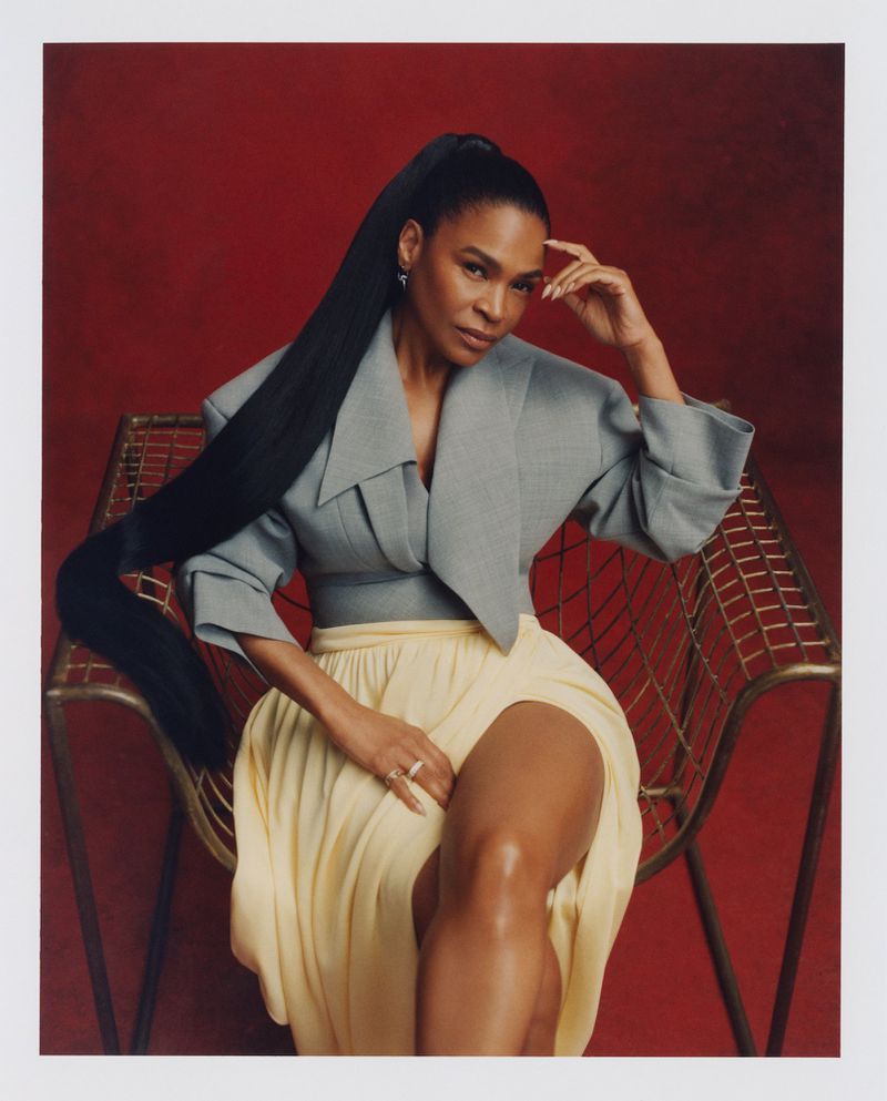 Adult female seated in a gold chair, wearing a gray crop top, and a yellow skirt, with a red background and studio lighting.