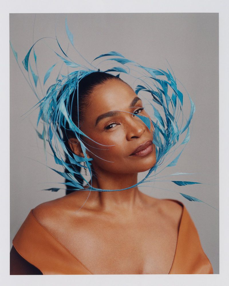 Portrait of an adult female with tan skin, wearing a sleeveless caramel-colored dress and a large, elaborate blue feathered headpiece.