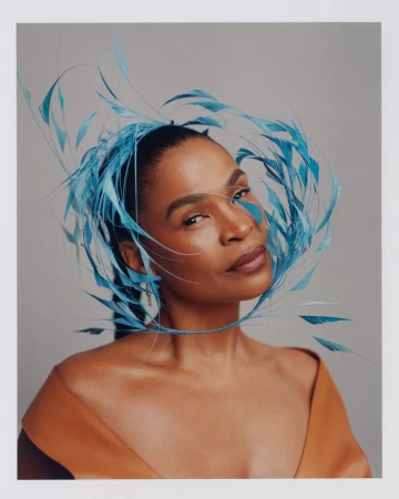 Portrait of an adult female with tan skin, wearing a sleeveless caramel-colored dress and a large, elaborate blue feathered headpiece.