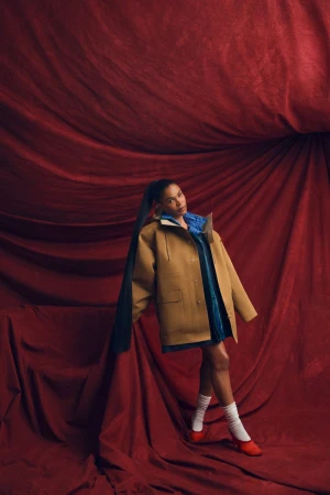 Woman in tan coat with long ponytail poses in front of draped red fabric in a fashion-style portrait with red shoes and white socks.