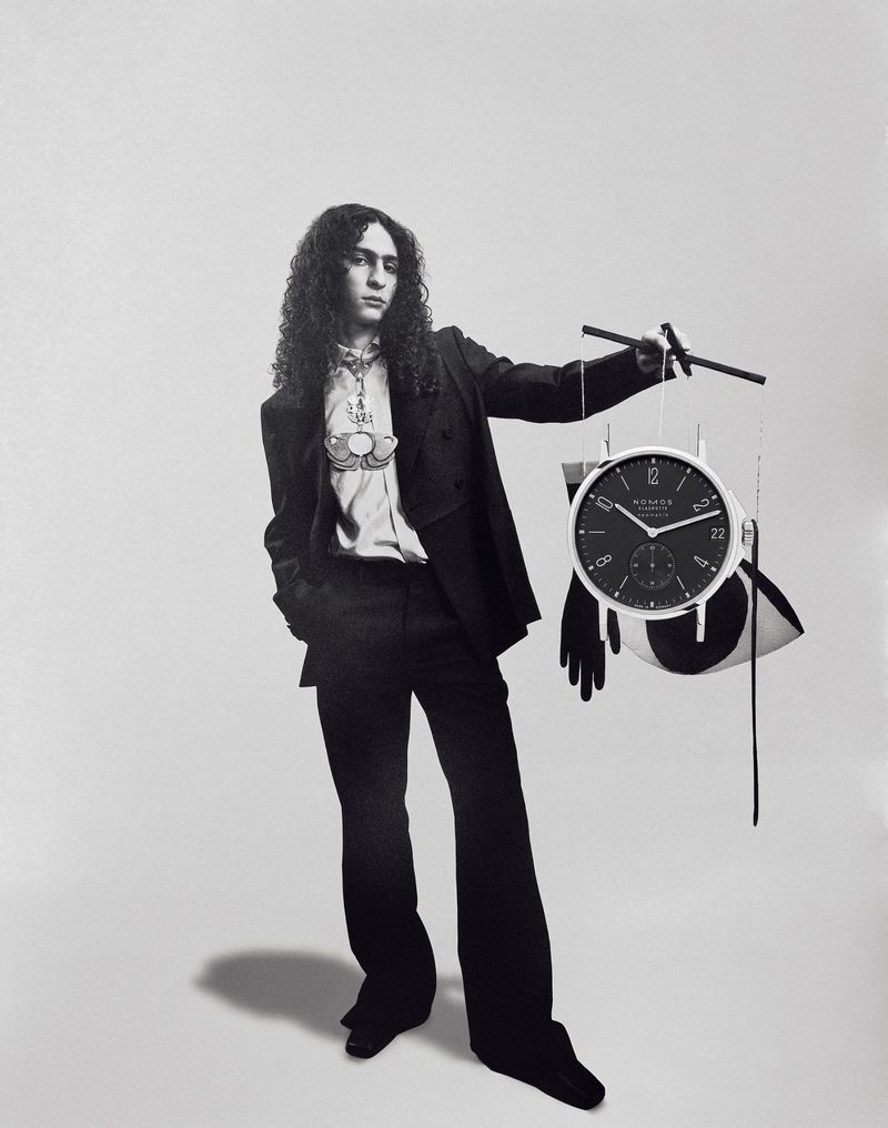 Black and white portrait of a stylish person with curly hair, wearing a suit, and posing with a clock and abstract elements.