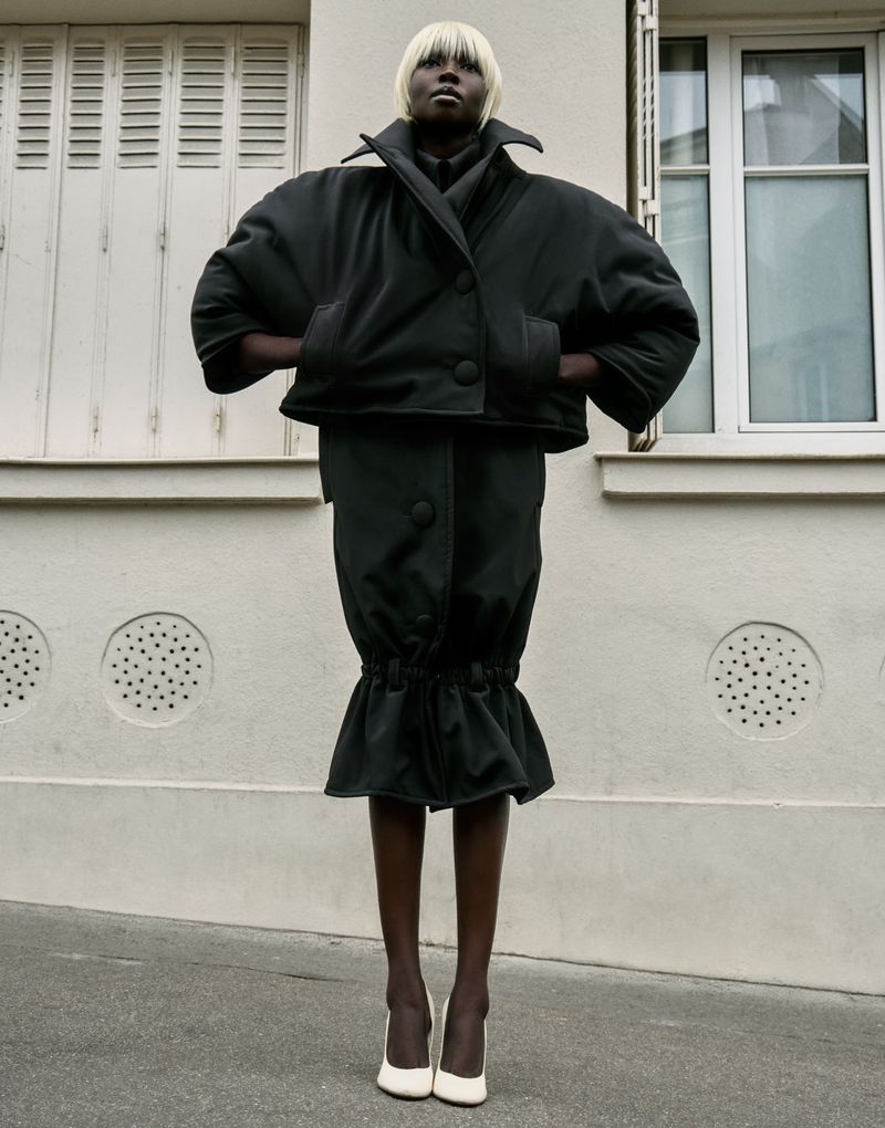 A dark-skinned woman with blonde hair and wearing a black jacket and skirt poses in front of a building with white shutters and walls.