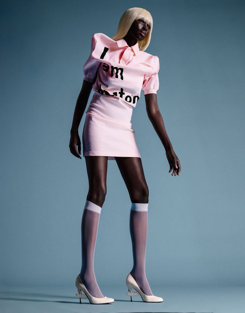 Model Ajok Madel wearing a light pink polo mini skirt set, blonde wig, and white heels against a blue seamless background.