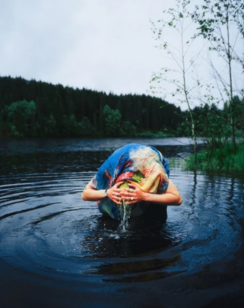 A person stands waist-deep in a lake with water cascading from their face, wearing a multicolored garment, surrounded by a forest.