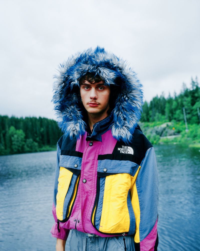 A young adult male poses in a colorful The North Face hooded jacket outdoors near a lake on a cloudy day, creating a lifestyle shot.