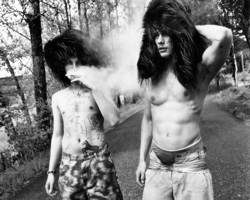 Black and white image of two bare-chested adult men with distinct hairstyles posing outside along a narrow road surrounded by trees.