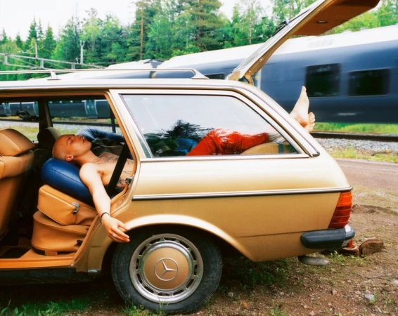 Bald adult lying inside of a gold car in an outdoor setting, seen next to a moving train and trees in the background.