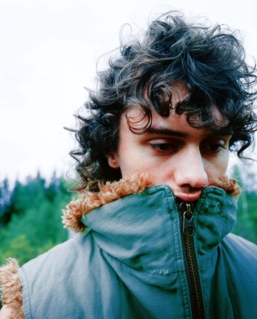 Portrait of a young adult male with curly hair wearing a teal vest with a fur collar standing outdoors in a forest landscape.