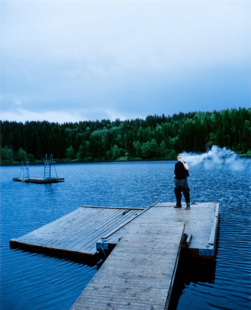 A person stands on a wooden dock by a blue lake with vapor emerging from their mouth, surrounded by a dark green forest under an overcast sky.