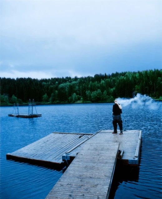 A person stands on a wooden dock by a blue lake with vapor emerging from their mouth, surrounded by a dark green forest under an overcast sky.