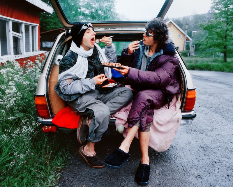 Two adults sit in the open trunk of a car, enjoying food with chopsticks. The setting is outdoors near a building and lush greenery.