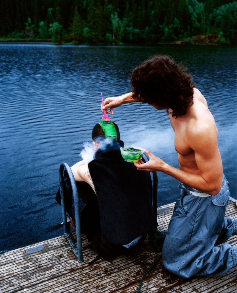 Two topless adult males on a wooden pier near a lake; one sits in a chair, receiving a treatment of smoking herbs from the other.
