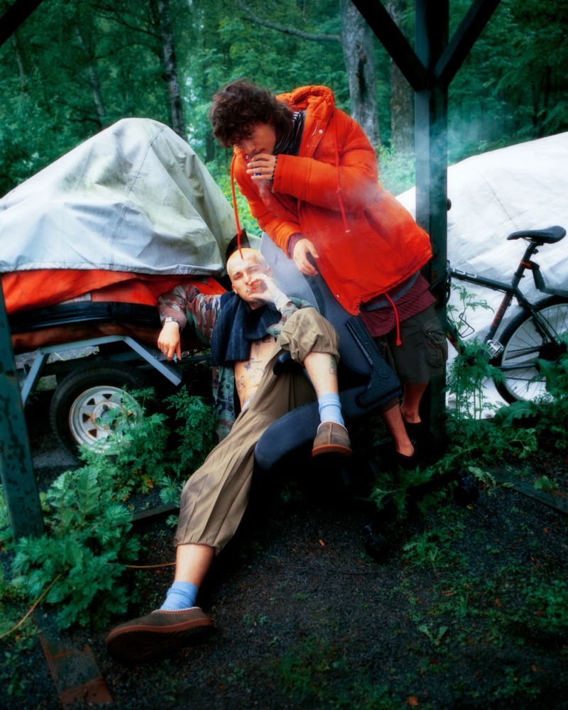 Two people in an outdoor setting, one seated and one standing, with a trailer and bike visible in the background. Lush greenery surrounds them.