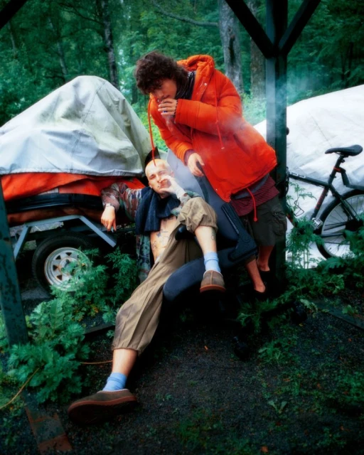 Two people in an outdoor setting, one seated and one standing, with a trailer and bike visible in the background.  Lush greenery surrounds them.